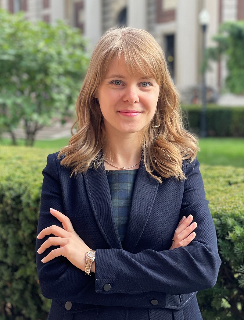 Headshot for Tatyana Avilova. Young white woman with blond hair, standing with her arms crossed on a college campus, looking at the camera. 