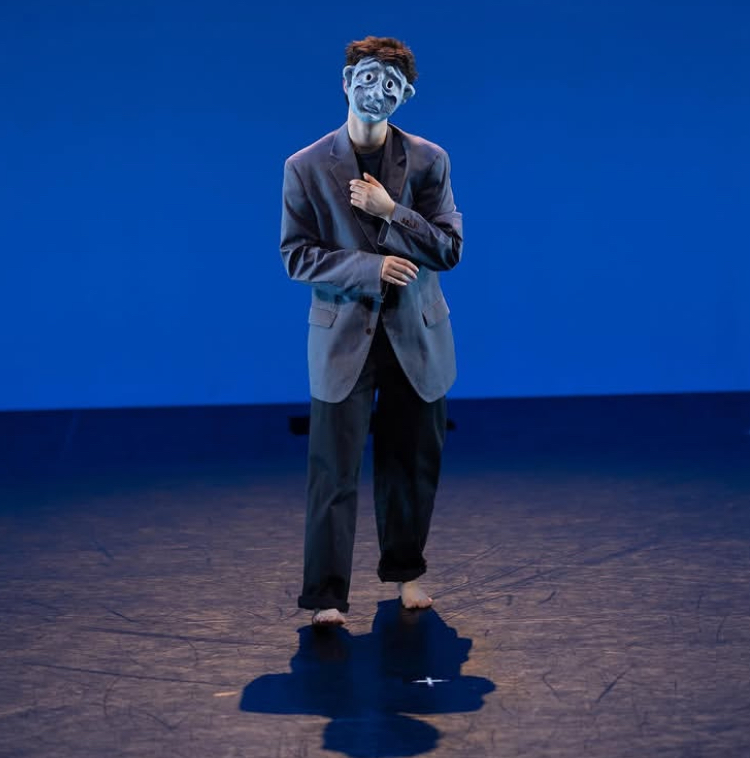 Person on suit on stage, wearing sad mask with big eyes